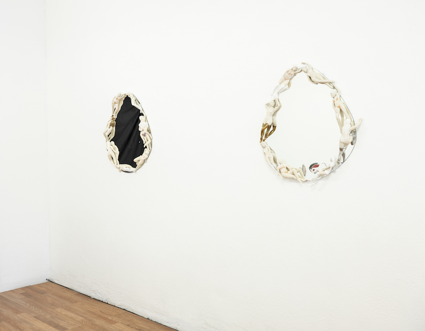 two wall mirrors handing on a white wall. In an egg shape surrounded by female figures in undergarments