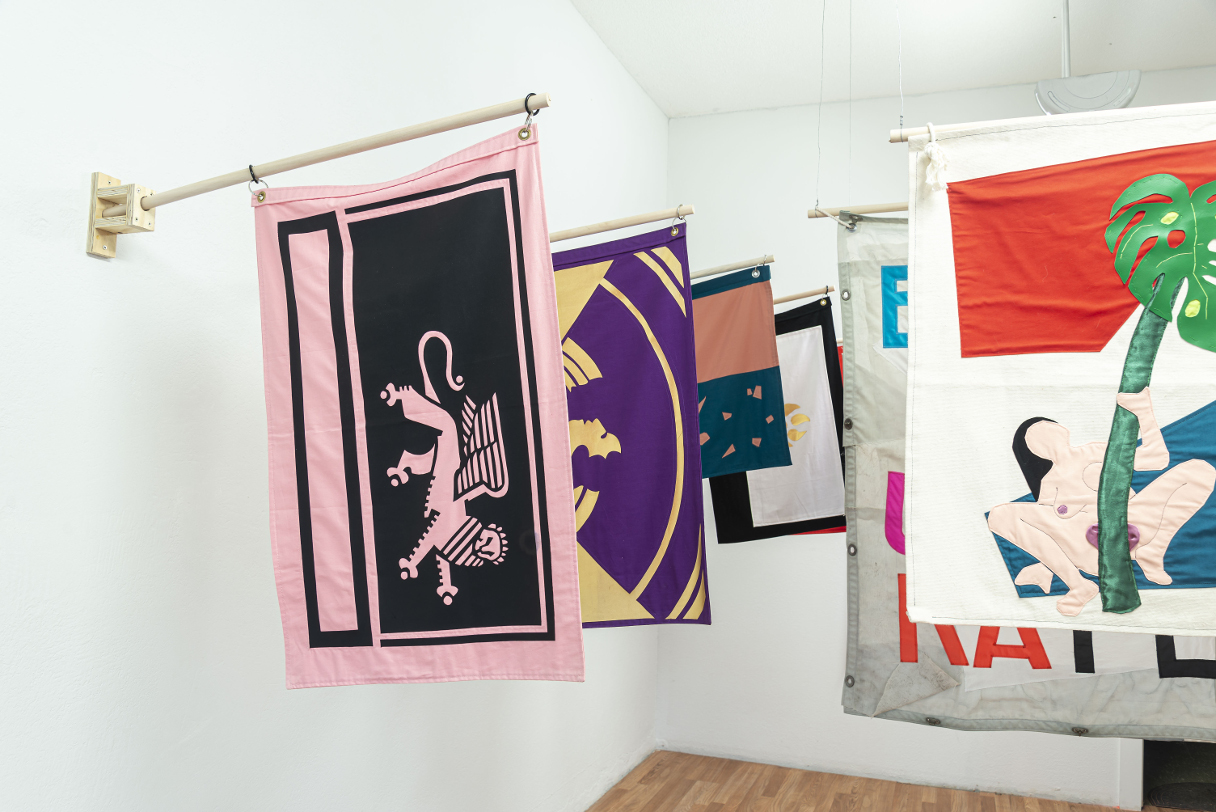 installation view: colorful flags hang in a clean well lit space with wooden floors and white walls